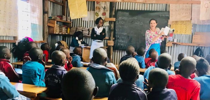 A woman stands in front of a chalkboard in front of a large classroom filled with children