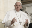 Pope Francis speaks while gesturing with his hands at a microphone, wearing white papal vestments and a cross pendant.