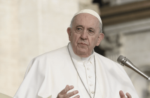 Pope Francis speaks while gesturing with his hands at a microphone, wearing white papal vestments and a cross pendant.