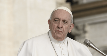 Pope Francis speaks while gesturing with his hands at a microphone, wearing white papal vestments and a cross pendant.