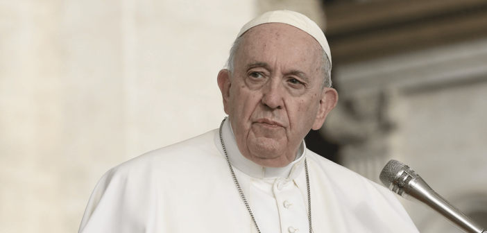 Pope Francis speaks while gesturing with his hands at a microphone, wearing white papal vestments and a cross pendant.