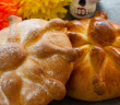 Two traditional pan de muerto loaves, a Mexican sweet bread made for Día de los Muertos. The round breads are decorated with bone-shaped dough pieces and dusted with sugar. One loaf has a golden-brown glaze, while the other is lightly covered with powdered sugar. In the background are bright orange and yellow paper marigolds and a small sugar skull decoration, set on a dark stone surface.